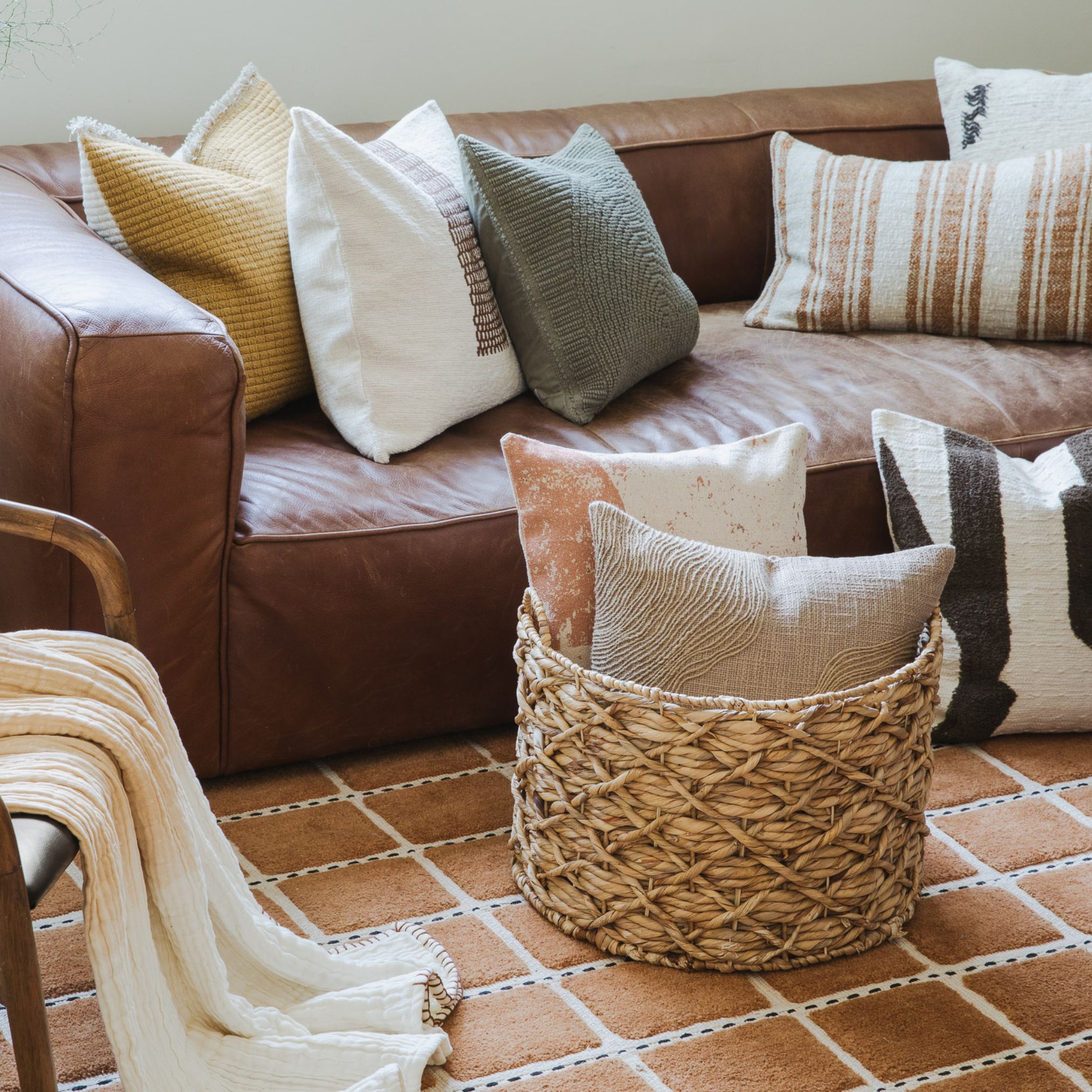 Brown leather sectional with assorted neutral throw pillows, woven basket, and beige blanket on a terracotta tile rug.