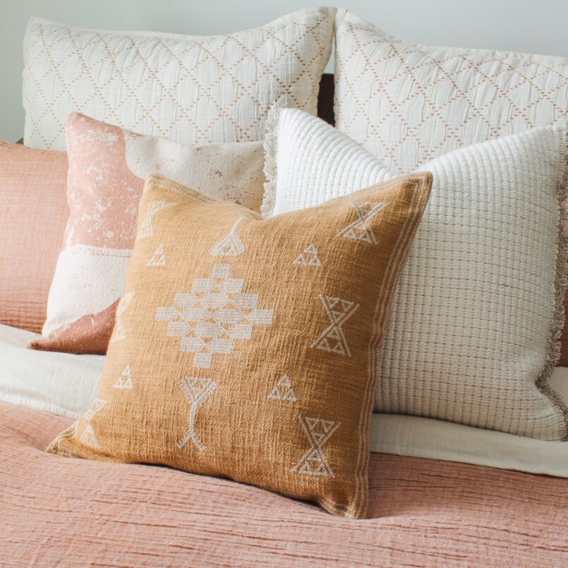 Burnt orange decorative pillow with white embroidery on a pink bed with white accent pillows