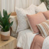 Soft pink bedding with striped and solid pillows, rust throw blanket, and small plant on bedside table