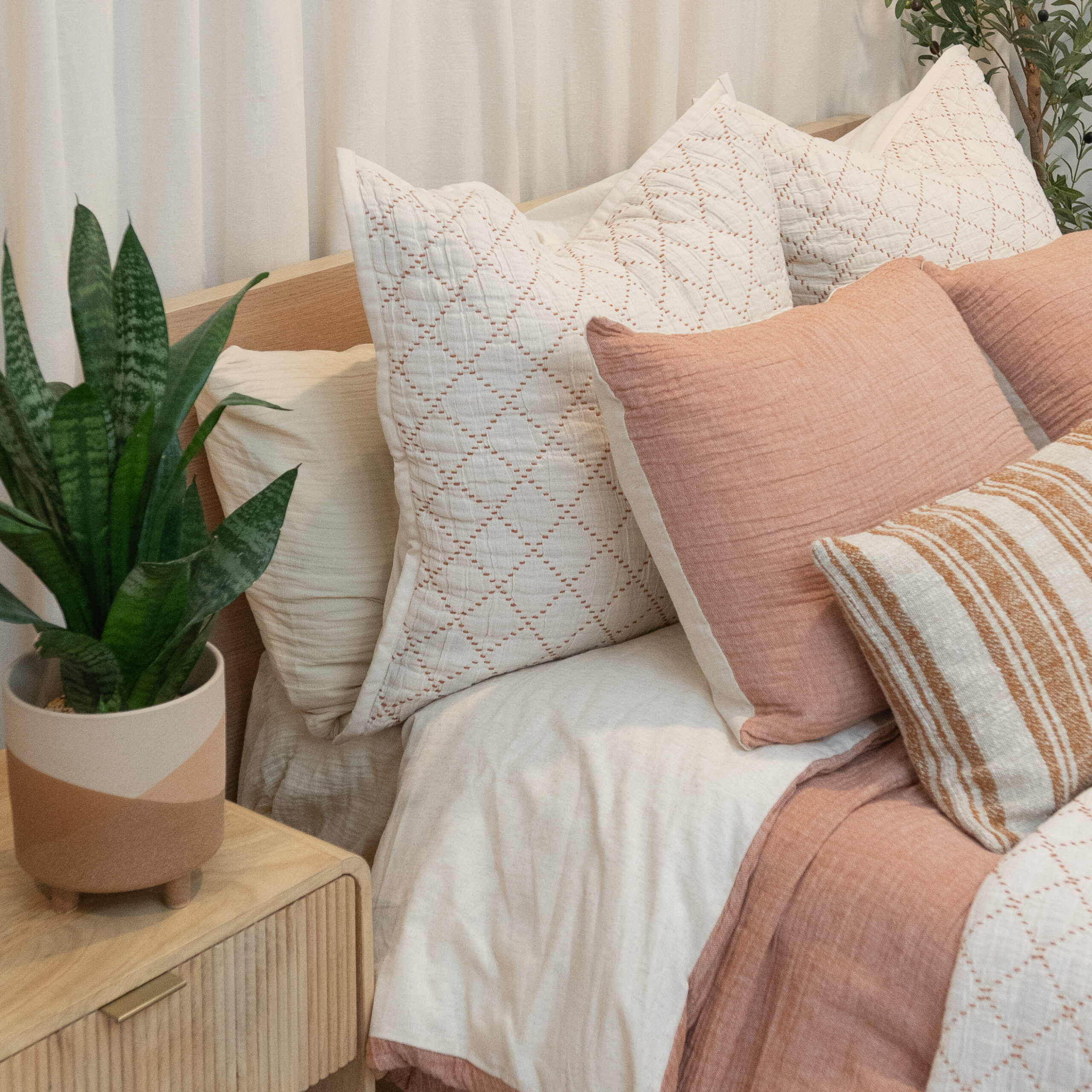 Soft pink bedding with striped and solid pillows, rust throw blanket, and small plant on bedside table