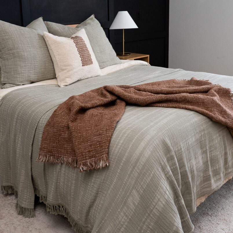 Light-colored bedding with a dark accent pillow and rust throw blanket in a softly lit room