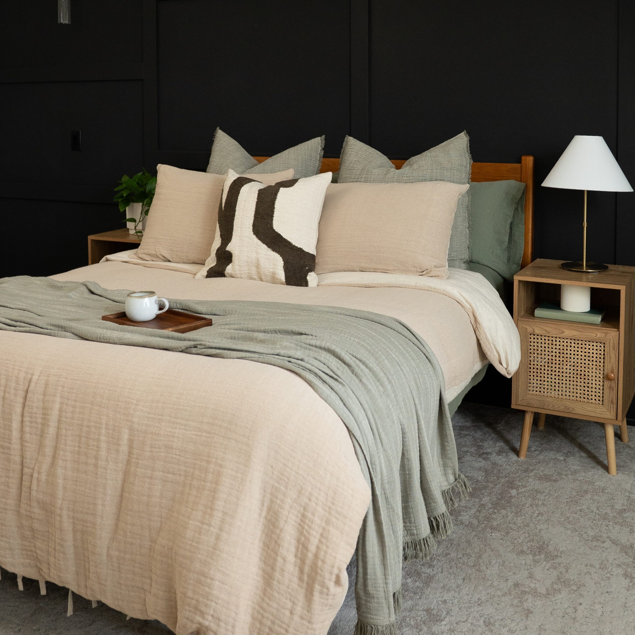 Beige bed with textured pillows and rust-colored blanket in front of a black wall and framed art