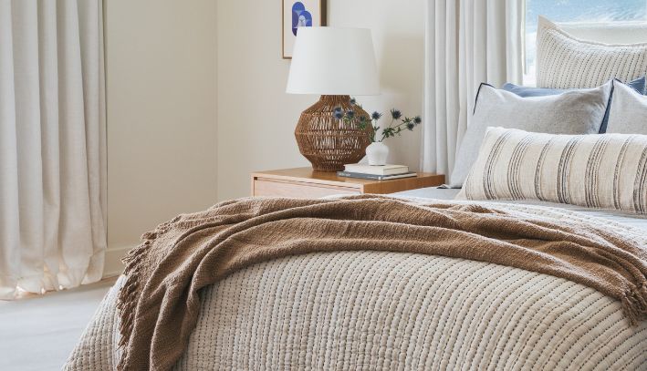 Neutral bedding set with a rust-colored throw blanket on a bed with a wooden headboard