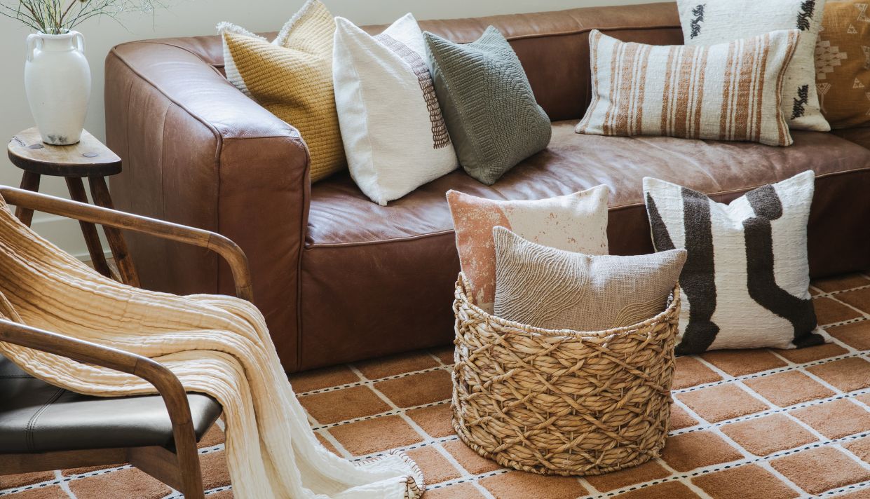 Brown leather couches with layered pillows and blankets, woven basket, and a wooden coffee table in a bright living room