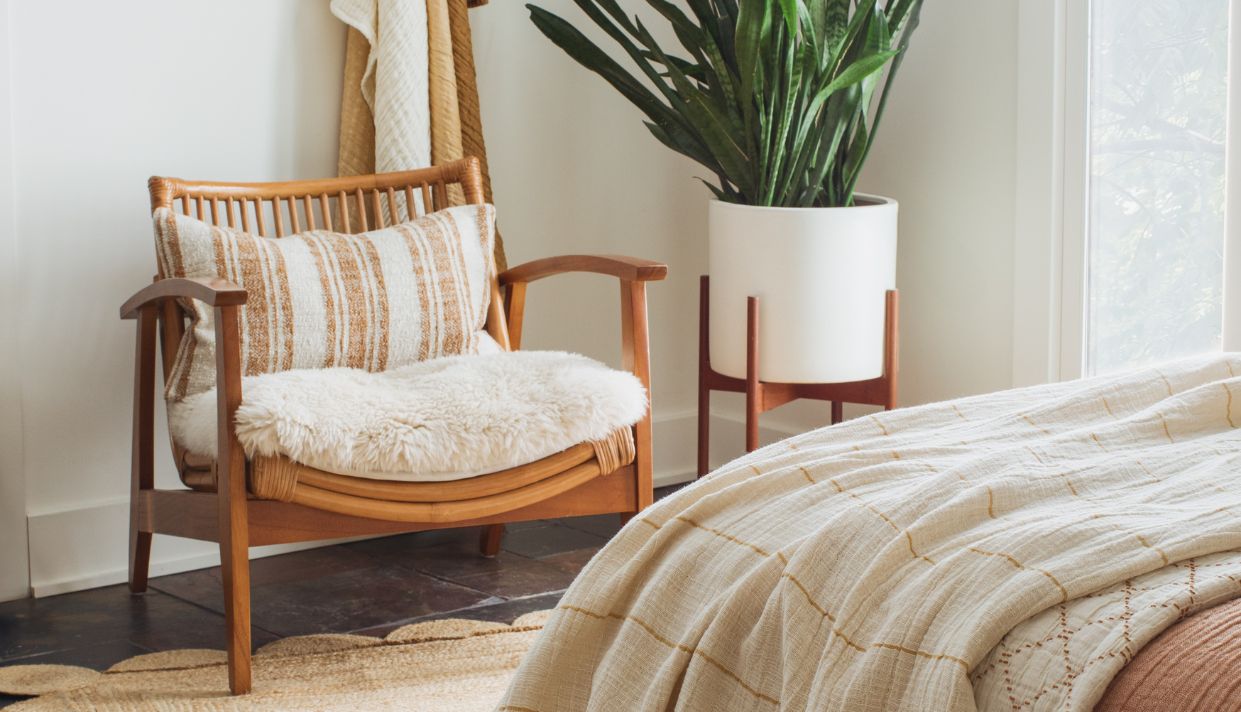 Beige armchair with wooden frame, layered pillows, and a plant in the background